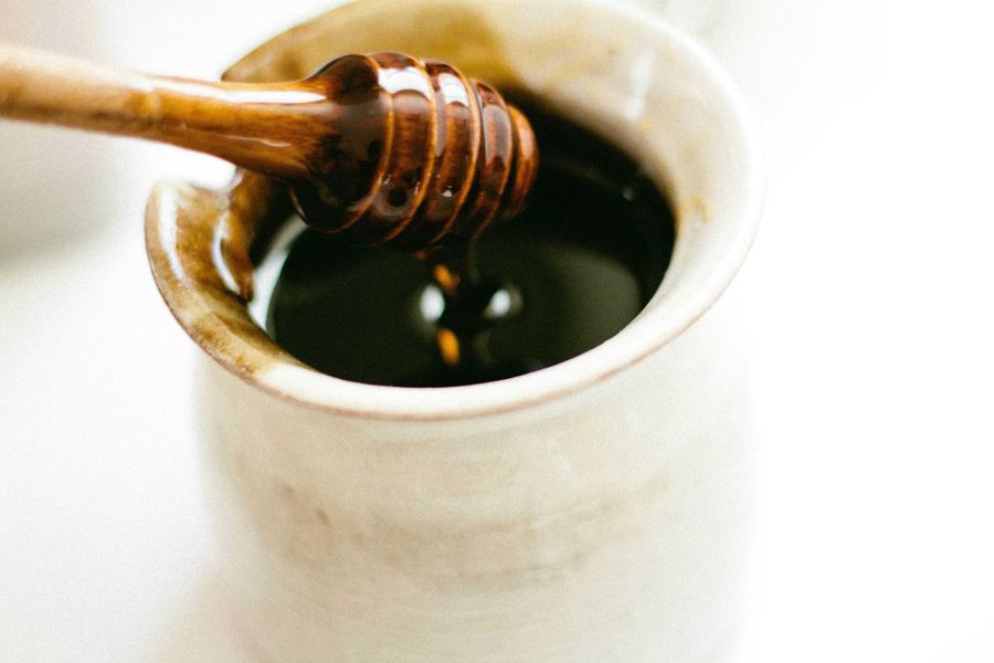 A wooden honey dipper resting in a small stoneware pot of dark honey