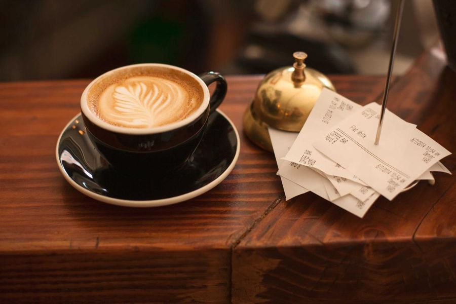 A flat white with leaf latte art on a dark saucer beside a brass service bell on a wooden bar